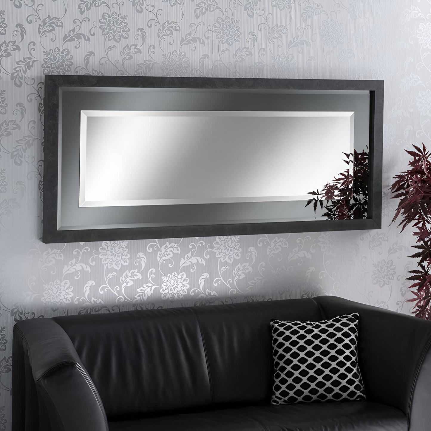Dark grey framed rectangular mirror on a wall with floral wallpaper, next to a black leather couch.