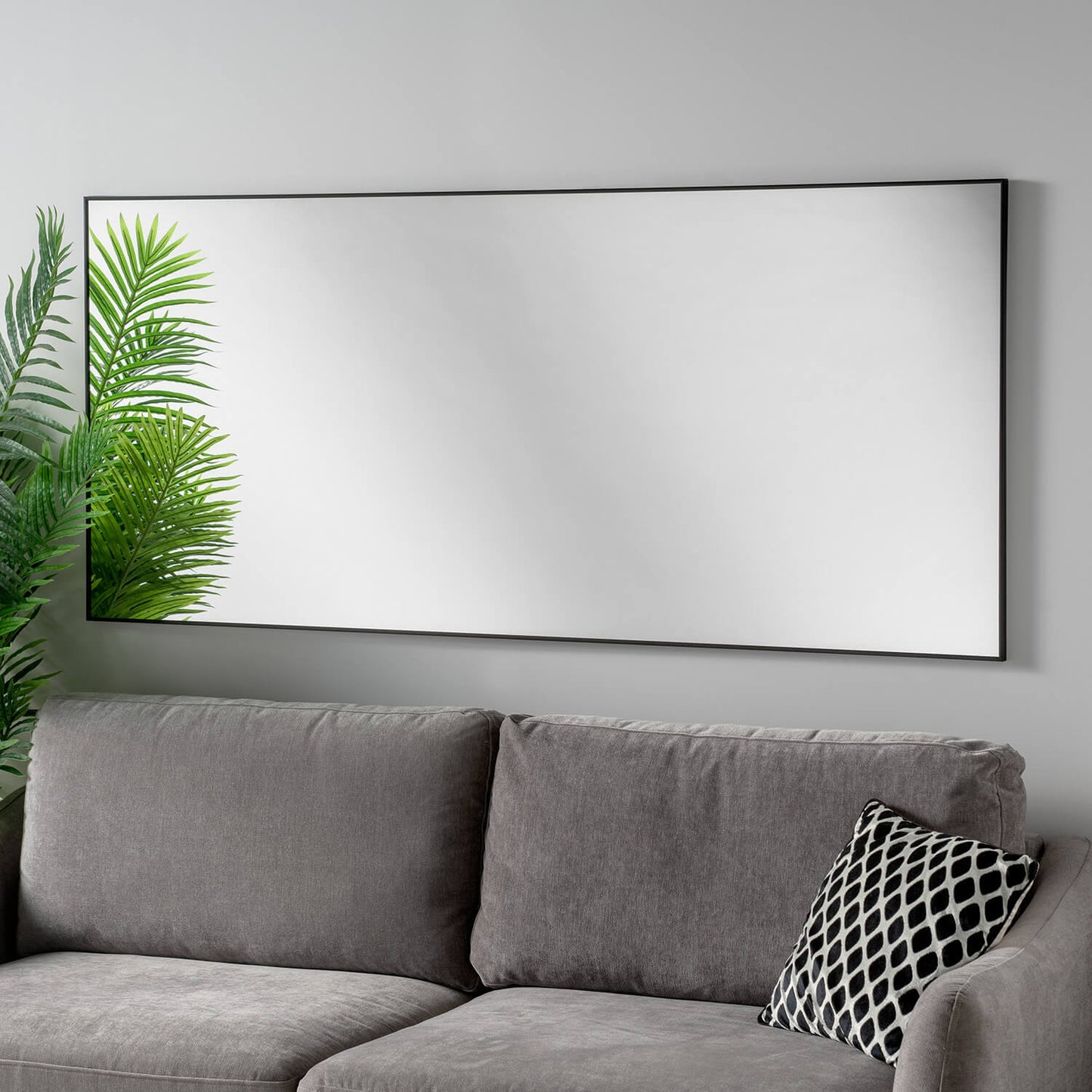 Large black framed mirror on a light grey wall, next to a grey couch with a patterned throw pillow, and a potted plant.