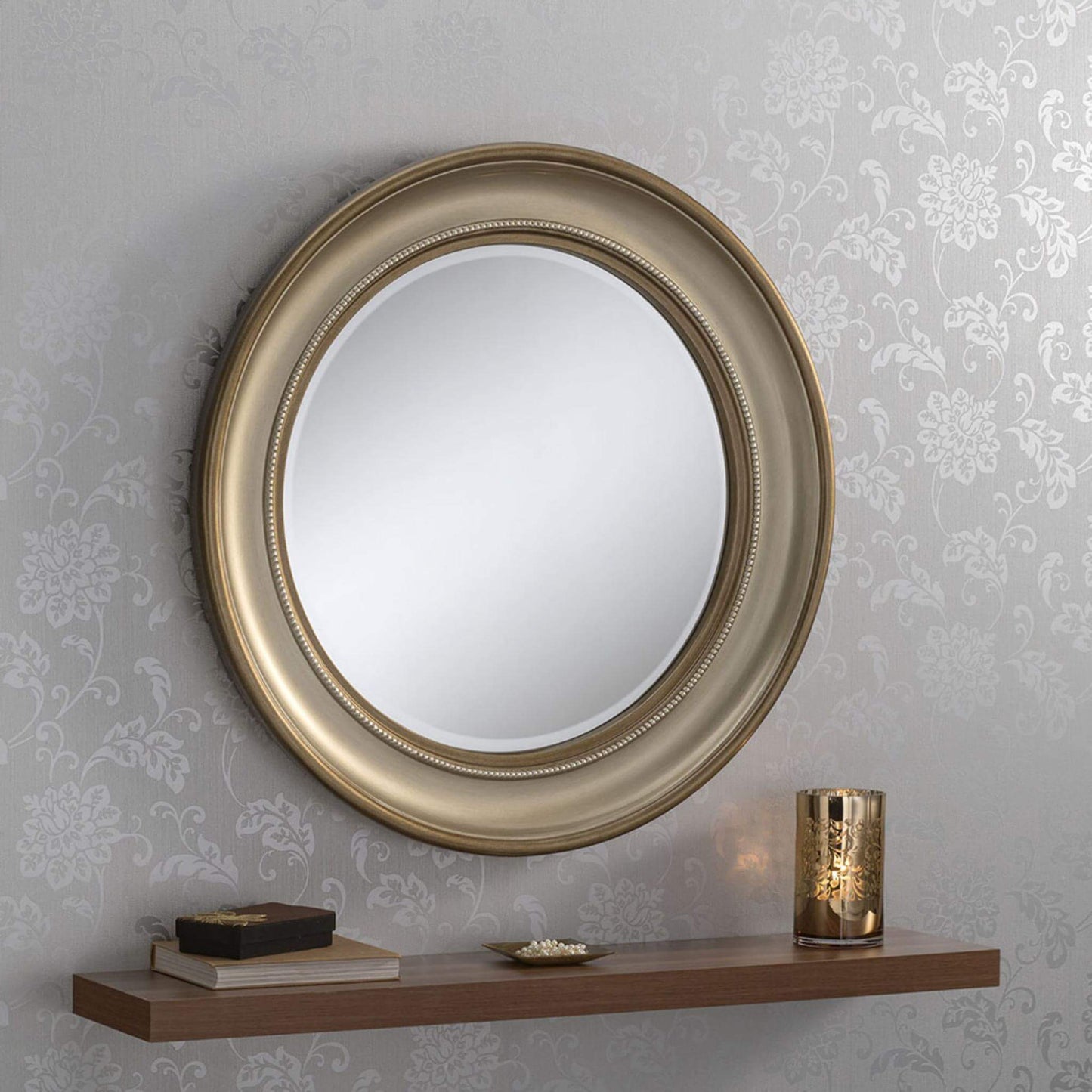 Circular bronze-framed mirror surrounded by beadlike detail, hung on a wall with floral wallpaper, and a shelf below displaying decorative items.