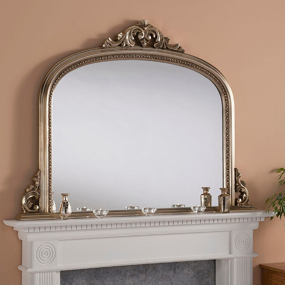 Antique-style, arched, silver-toned mirror mounted on a white mantelpiece with decorative accents.