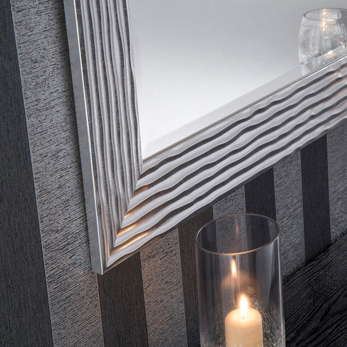 Silver-framed rectangular mirror with wavy design, against a dark gray striped wall, and a glass candle holder.
