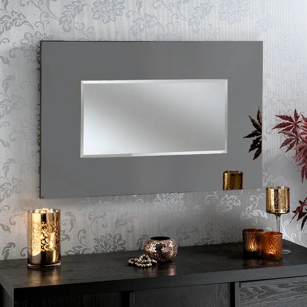 A rectangular wall mirror with a sleek black finish on a grey frame and a beveled edge. The mirror is mounted on a wall with a floral-patterned wallpaper. Below the mirror, there is a dark wooden surface with decorative items, including a gold candle holder, a small metallic bowl with beads, and two lit candles in glass holders