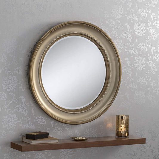 Circular bronze-framed mirror  surrounded by beadlike detail, hung on a wall with floral wallpaper, and a shelf below displaying decorative items.