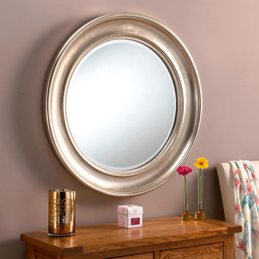 Large round silver-framed mirror hangs on a mauve wall, atop a wooden vanity with decorative items.